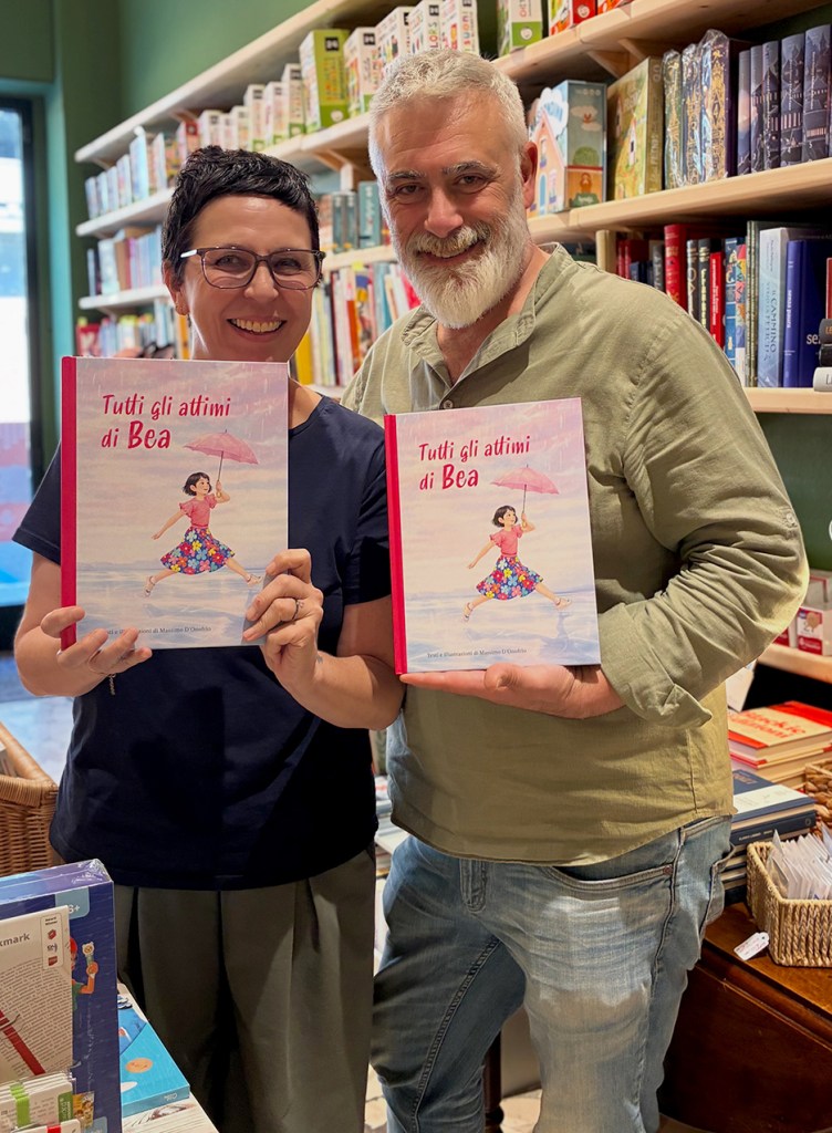 Due persone sorridenti in una libreria, entrambe tengono in mano il libro 'Tutti gli attimi di Bea'. Sullo sfondo si vedono scaffali pieni di libri.
