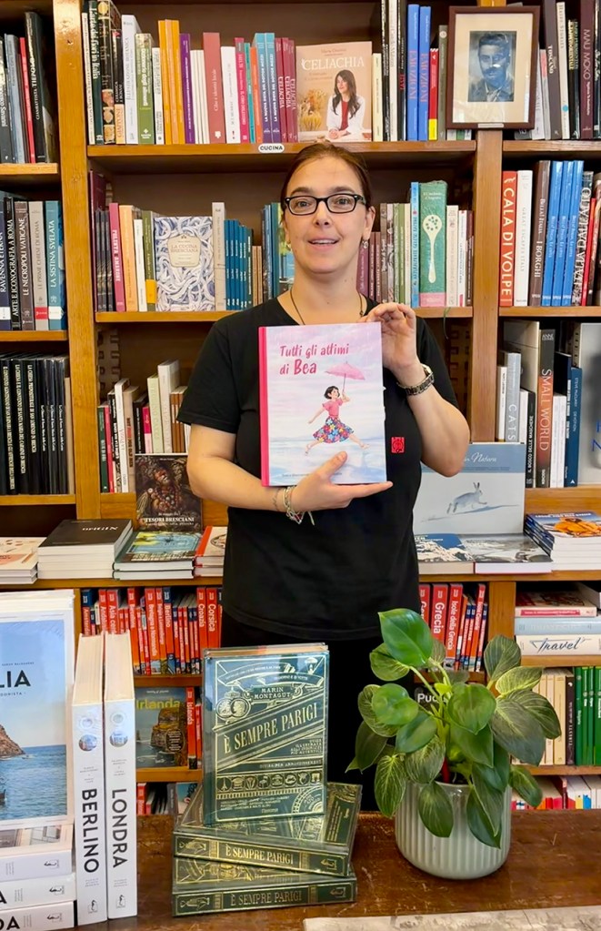 Una donna in una libreria tiene in mano il libro 'Tutti gli attimi di Bea', con una libreria di sfondo che contiene vari libri e piante decorative.