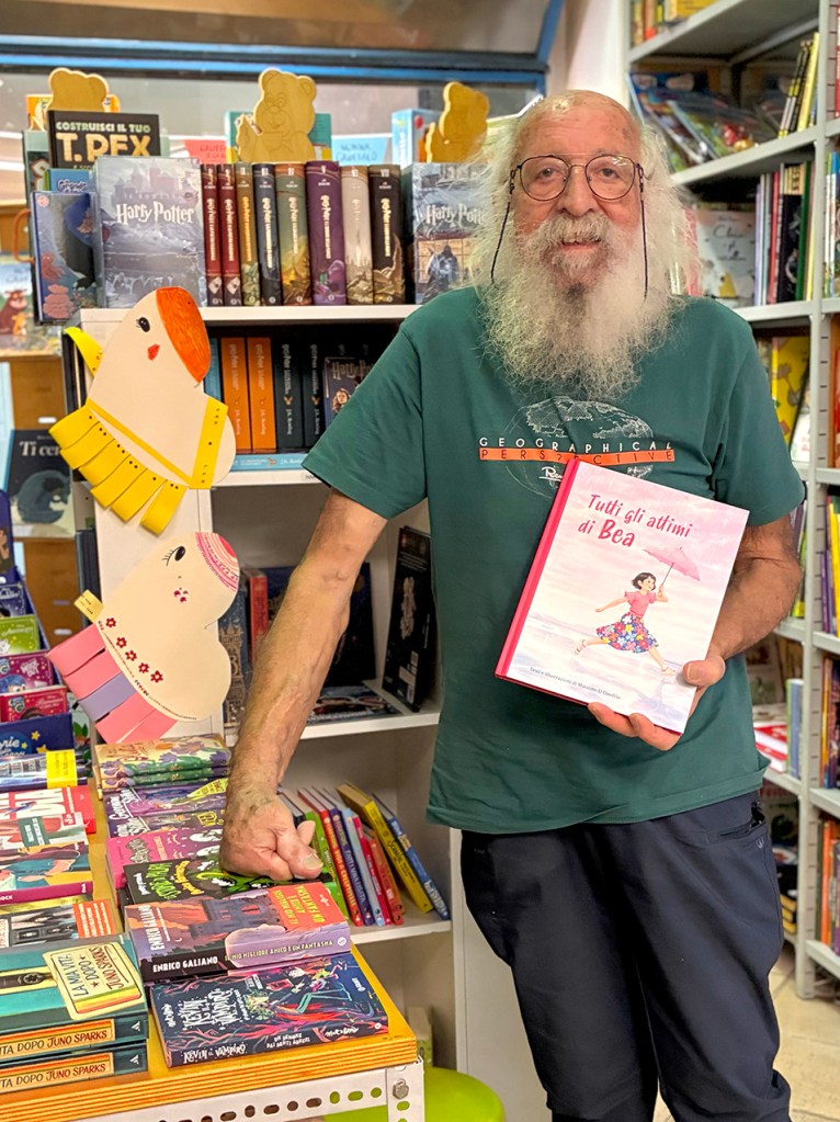 Un uomo anziano con barba bianca e occhiali tiene in mano un libro intitolato 'Tutti gli attimi di Bea' in una libreria, circondato da libri e decorazioni colorate.