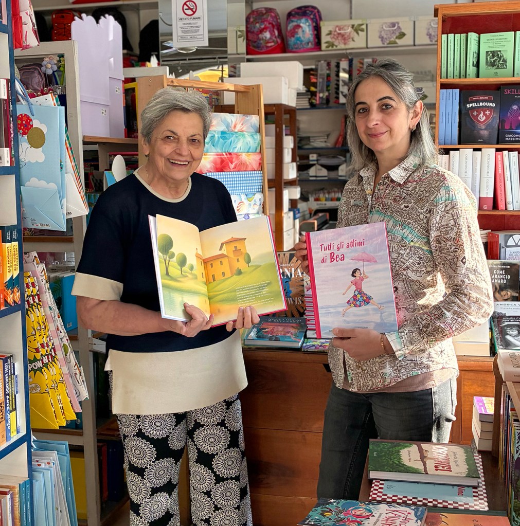 Due donne sorridenti in una libreria, ciascuna tiene un libro illustrato. Una mostra un libro con un paesaggio, mentre l'altra tiene un libro intitolato 'Tutti gli attimi di Bea'.
