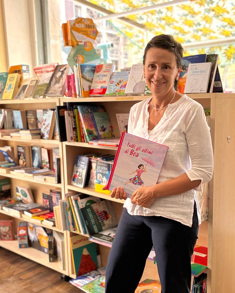Una donna sorridente tiene un libro intitolato 'Tutti gli attimi di Bea' in una libreria, con uno scaffale di libri sullo sfondo.