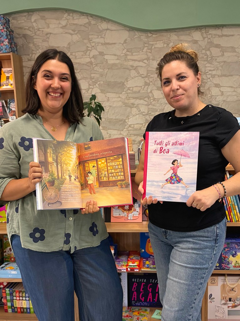 Due donne sorridenti in una libreria mostrano i libri infantili "Una favolosa scoperta" e "Tutti gli attimi di Bea".