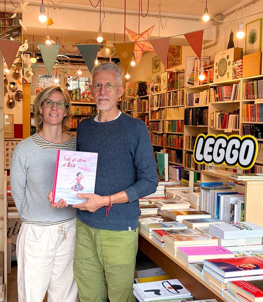Due persone in una libreria, sorridenti, tengono in mano un libro intitolato 'Tutti gli altini di Bea', circondati da scaffali pieni di libri.