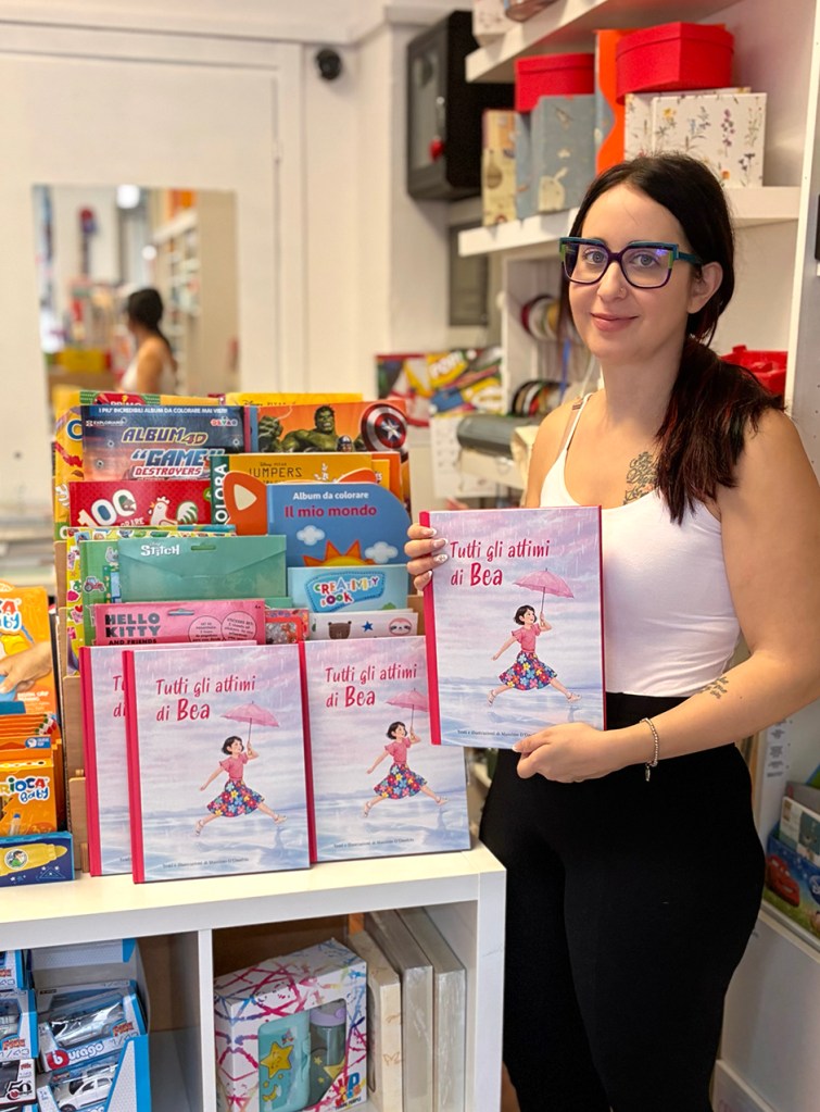 Una donna in una libreria tiene in mano un libro intitolato 'Tutti gli attimi di Bea', circondata da vari giochi e bambini all'interno di scaffali colorati.