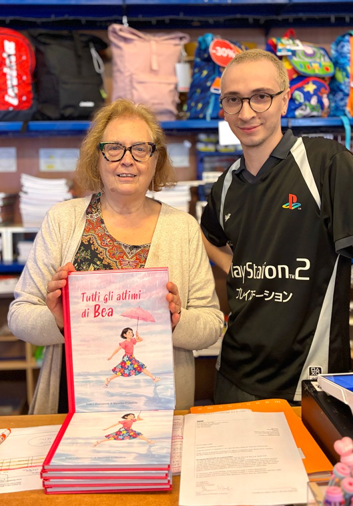 Due persone sorridenti mostrano un libro intitolato 'Tutti gli attimi di Bea' in un negozio, con zaini colorati sullo sfondo.