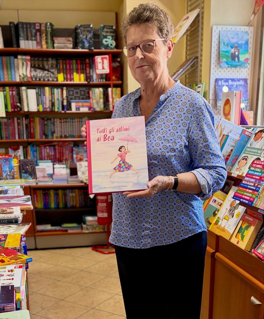 Una donna in una libreria tiene in mano un libro intitolato 'Tutti gli attimi di Bea', con uno sfondo di scaffali pieni di libri.