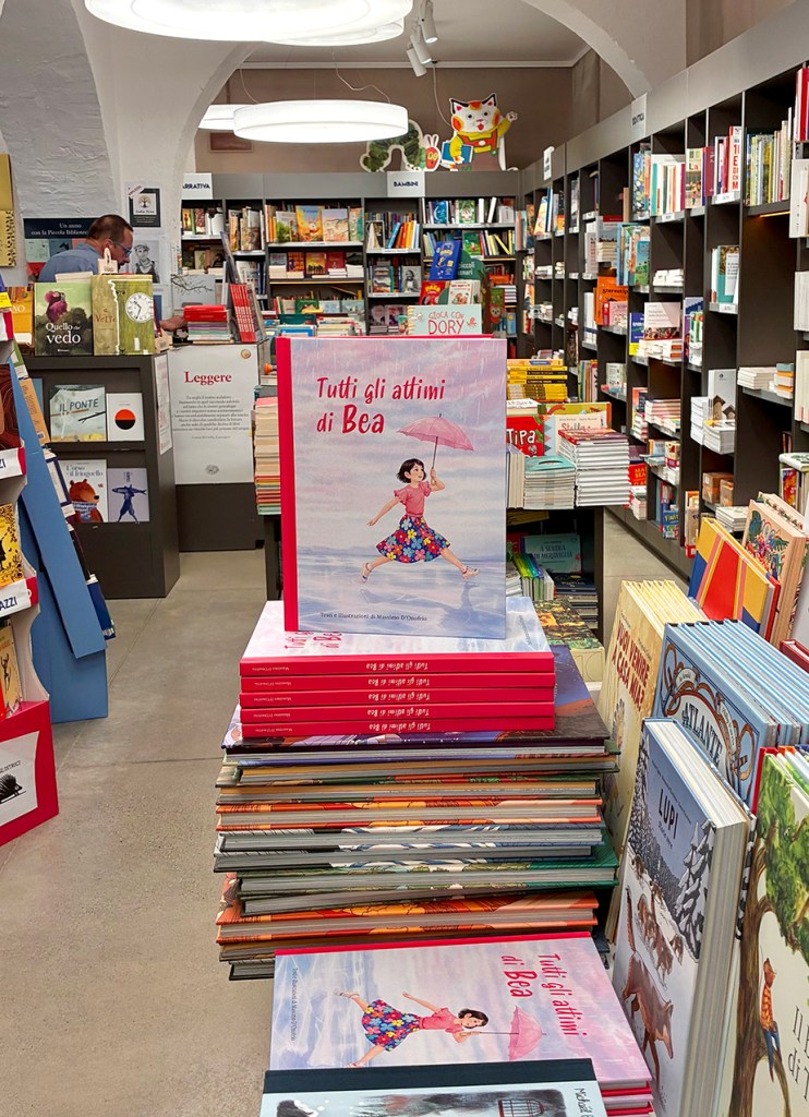 Un negozio di libri con scaffali pieni di volumi. In primo piano, un libro intitolato 'Tutti gli attimi di Bea' su una pila di libri.