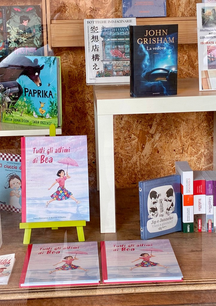Libreria con vari libri esposti, tra cui 'Tutti gli attimi di Bea' in primo piano, illustrato con una bambina che salta con un ombrello rosa.