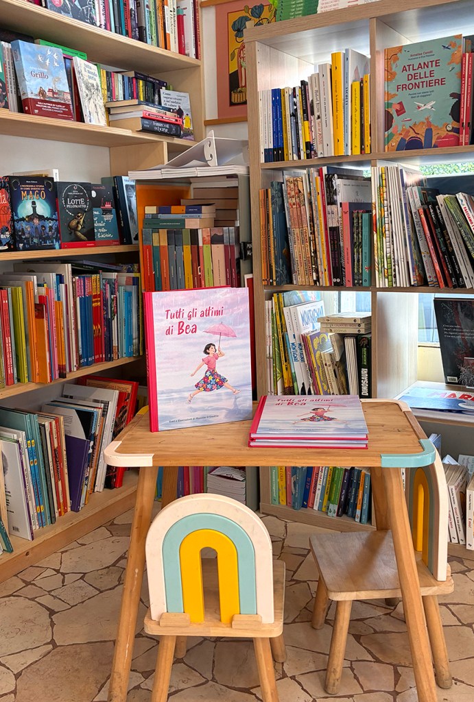 Libreria con scaffali pieni di libri colorati e un tavolo di legno con il libro 'Tutti gli attimi di Bea' in evidenza.
