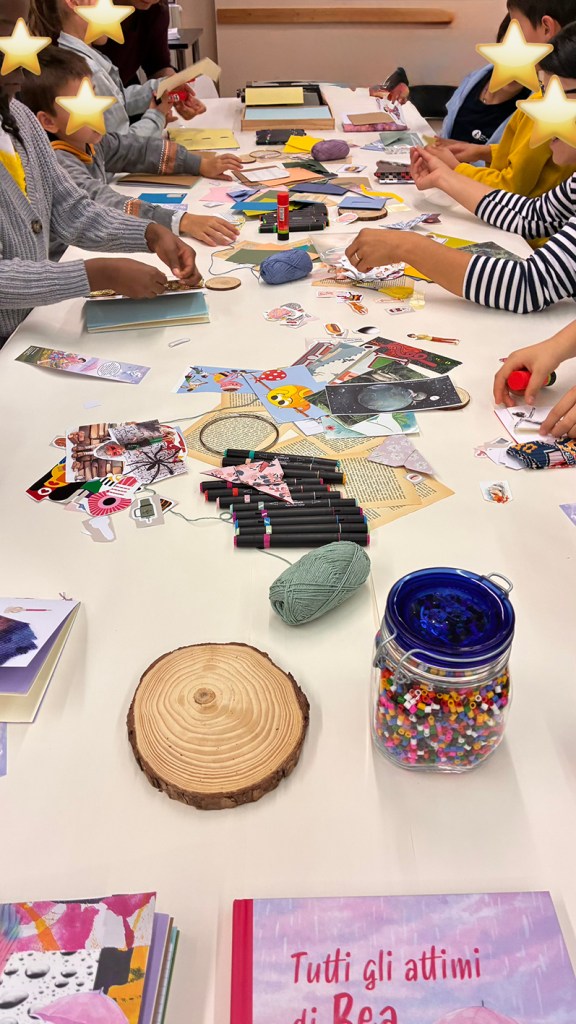 Un tavolo pieno di materiale creativo, con bambini e adulti che realizzano progetti artistici. Ci sono libri, forbici, colla, penne, ritagli di carta colorata e perline, insieme a gomitoli di lana e un disco di legno.