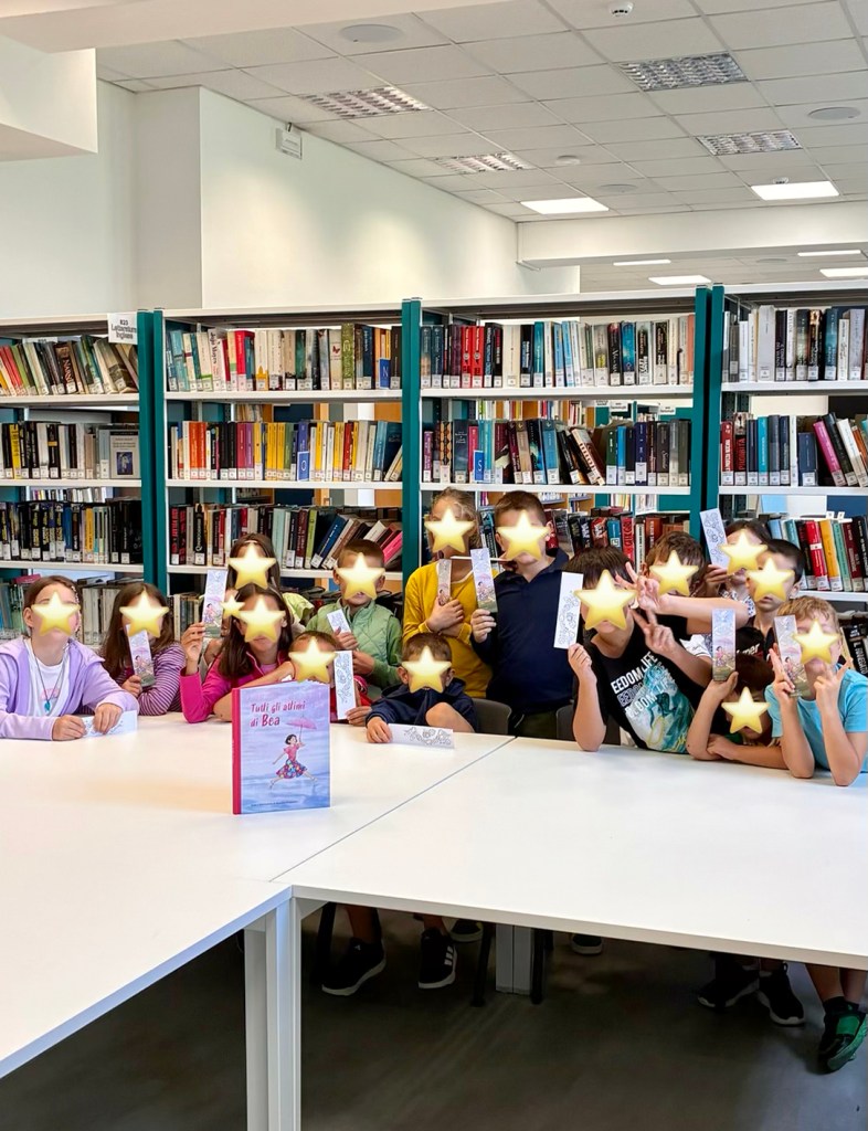 Gruppo di bambini in una biblioteca, che tengono dei segnalibri alzati davanti a un libro intitolato 'Tutti gli amici di Bea'.